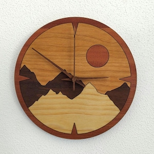 May include: A round wooden wall clock with a landscape design. The clock face features a mountain range silhouette, a sun, and wooden clock hands. The clock is made of various shades of brown wood.