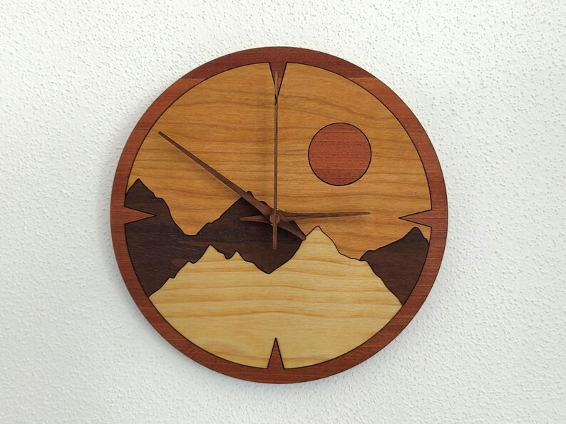 May include: A round wooden wall clock with a landscape design. The clock face features a mountain range silhouette, a sun, and wooden clock hands. The clock is made of various shades of brown wood.