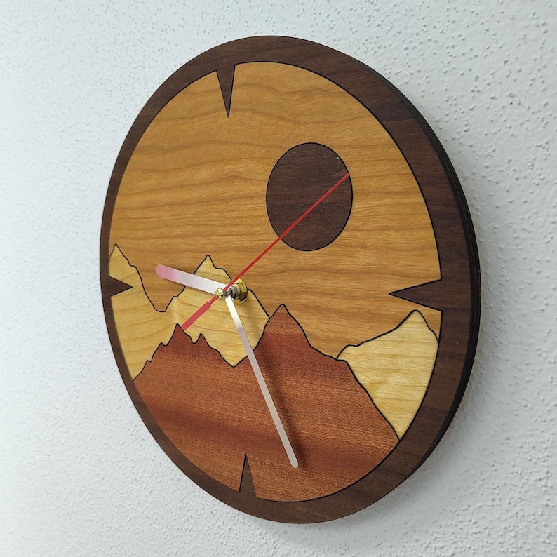 May include: A round wooden clock with a mountain landscape design. The clock face features a sun and layered mountains in shades of brown and tan. The clock hands are silver and red, with a dark brown wooden frame.