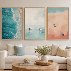 May include: A set of three framed wall art prints featuring coastal scenes. The left print shows an aerial view of turquoise ocean waves hitting a sandy beach. The center print depicts two figures with surfboards in shallow water. The right print shows a sandy beach with two small umbrellas.