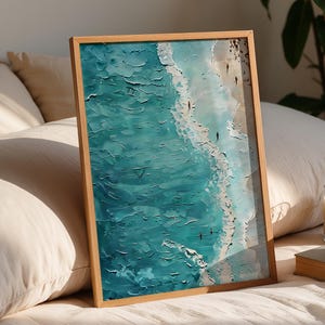 May include: A framed art print depicting a coastal scene with turquoise water and a sandy beach. The artwork is in a wooden frame and rests on a bed with pillows. A candle and a cup of coffee are also visible.
