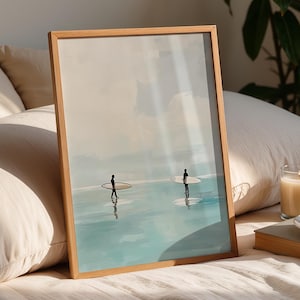 Puede incluir: Una impresión enmarcada representa a dos personas con tablas de surf en una escena costera serena. La obra de arte, en un marco de madera clara, presenta una paleta de colores suaves de azules y beige. Una vela encendida, una taza de café y un libro están en una cama con almohadas.