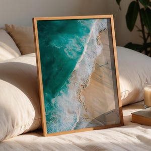 May include: A framed print of a turquoise ocean wave crashing on a sandy beach. The print has a natural wood frame and is displayed on a bed with pillows. A candle, a cup of coffee, and a book are also on the bed.