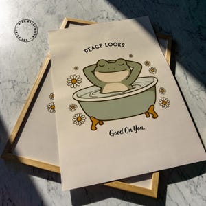 Puede incluir: Una impresi&oacute;n art&iacute;stica enmarcada con una rana de dibujos animados en una ba&ntilde;era. La rana es verde y beige, con los brazos detr&aacute;s de la cabeza. La impresi&oacute;n incluye el texto "Peace Looks Good On You" e ilustraciones de margaritas.