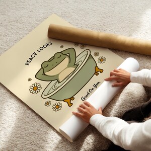 Puede incluir: Un p&oacute;ster beige con una rana de dibujos animados relaj&aacute;ndose en una ba&ntilde;era, rodeada de margaritas. El texto "Peace Looks Good On You" est&aacute; impreso en el p&oacute;ster. Tambi&eacute;n se ven un p&oacute;ster enrollado y un tubo marr&oacute;n.