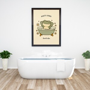 Puede incluir: Un cuadro enmarcado con un dibujo de una rana de dibujos animados relaj&aacute;ndose en una ba&ntilde;era, con el texto "Peace Looks Good On You". La obra de arte est&aacute; sobre un fondo crema, rodeada de margaritas. El ba&ntilde;o tiene una ba&ntilde;era blanca y plantas.