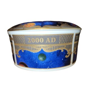 May include: A ceramic trinket box with a lid, featuring a celestial design. The box has a white base with a blue and gold celestial pattern. The text "2000 AD" is displayed on the side. The lid has a similar design with planets.