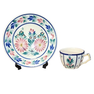 Könnte beinhalten: Eine weiße Teetasse und Untertasse aus Keramik mit einem floralen Design in Rosa-, Blau- und Grüntönen. Die Tasse hat einen Henkel und die Untertasse einen gewellten Rand.