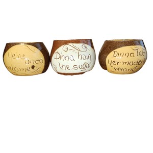 May include: Three brown ceramic mugs with white lettering. The mugs have the following text: "Here once welcome", "Dinna-han the sugar", and "Dinna let yer modest wrangl".