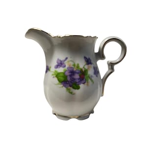 May include: A white porcelain creamer with a floral violet design. The creamer has a gold trim around the rim and handle. The handle is a loop shape. The creamer is approximately 5 inches tall.