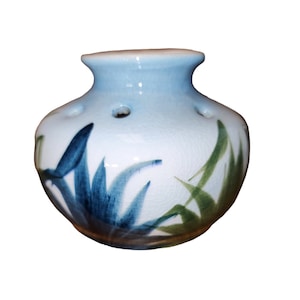 May include: A white ceramic vase with blue and green floral designs. The vase has a round body and a small opening at the top. There are several small holes around the opening.