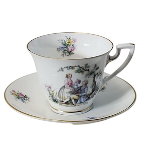 Puede incluir: Un juego de taza y platillo de té blanco con ribete dorado. La taza y el platillo presentan un patrón floral colorido y una escena de dos personas en un jardín.