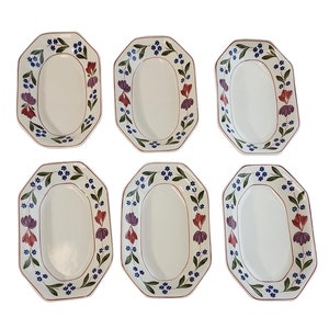 May include: Six white oval serving platters with a floral design. Each platter has a red rim and is decorated with purple, red, and blue flowers and green leaves. The platters are arranged on a white background.