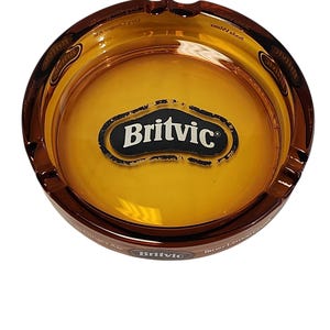 Può includere: Un posacenere rotondo in vetro color ambra con il logo Britvic al centro. Il bordo del posacenere presenta diverse tacche per appoggiare oggetti, e il lato esterno mostra il marchio Britvic insieme a del testo relativo al Ginger Ale.