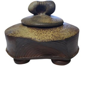 Pode incluir: Um pote de cerâmica artesanal com tampa. O pote é marrom escuro com um topo salpicado marrom claro e bege. A tampa tem um formato orgânico único. O pote fica sobre três pés pequenos.