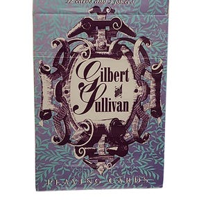 Può includere: Un mazzo di carte da gioco in una scatola rettangolare con sfondo viola chiaro e un motivo a foglie color ottanio. Al centro è presente un emblema viola scuro con il testo Gilbert and Sullivan. In alto si legge 52 cards and 3 jokers.