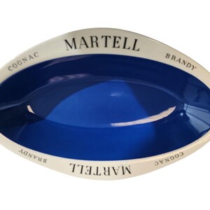 Può includere: Un piatto ovale in ceramica con interno blu profondo e bordo color crema. Il bordo presenta il testo nero MARTELL, COGNAC e BRANDY stampato ripetutamente lungo il perimetro. Il piatto ha una finitura liscia e lucida.