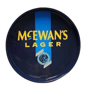 Può includere: Un vassoio da portata rotondo in metallo blu scuro con la scritta gialla MCEWAN'S LAGER. Una striscia verticale azzurra attraversa il centro, dietro un logo circolare che raffigura una persona di profilo con il testo BREWER SINCE 1856.