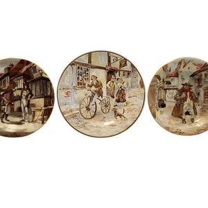 Può includere: Tre piatti circolari in ceramica con scene storiche dettagliate. Il piatto a sinistra mostra figure in una strada di paese. Il piatto centrale raffigura una persona in bicicletta con un cane. Il piatto a destra mostra una figura che cammina in una piazza.