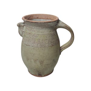 May include: A handmade, rustic ceramic pitcher with a handle and a small spout. The pitcher has a light green and brown speckled glaze, with a darker brown rim and base. The overall shape is rounded, with a wide body and a narrow neck.