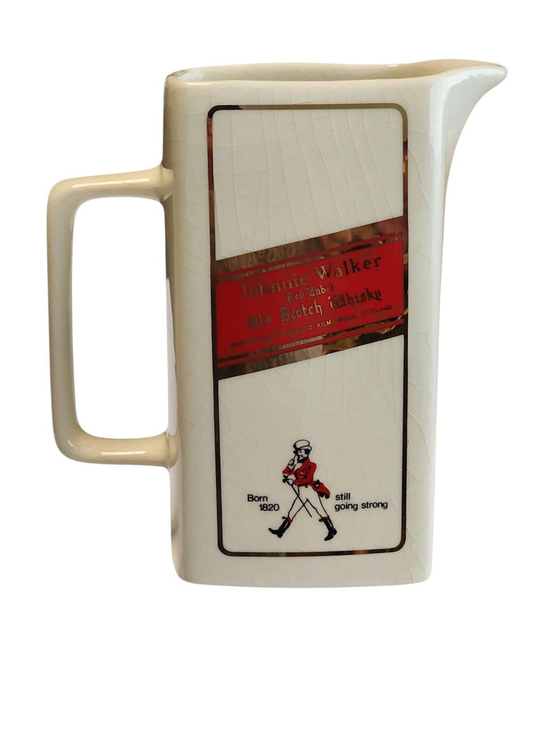 May include: Cream ceramic pitcher with a rectangular design. It features a red and gold label with the text "Johnnie Walker Red Label Scotch Whisky." Below the label is a graphic of a man in a red coat and the text "Born 1820 still going strong."