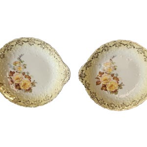 May include: Two vintage, off-white ceramic serving dishes with gold trim and handles. Each dish features a floral design with yellow and peach roses. The edges have a textured, scalloped design.