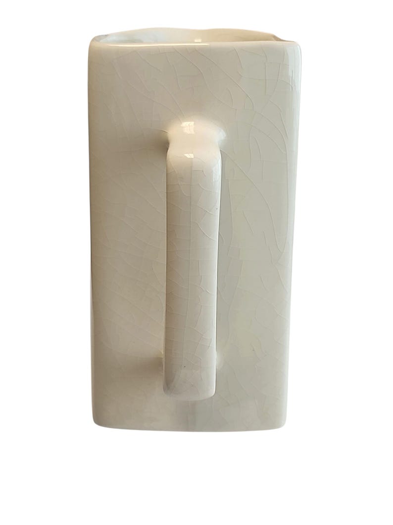 May include: Cream-coloured ceramic mug with a rectangular body and a curved handle. The mug has a crackle glaze finish, adding a textured appearance. The handle is smooth and rounded, contrasting with the textured body.