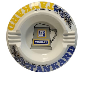 Può includere: Un posacenere in ceramica bianca con una grafica centrale di un boccale di birra grigio con il testo TANKARD. Il bordo mostra la parola TANKARD in grandi lettere gialle e blu, insieme a illustrazioni floreali blu e il testo WHITBREAD.