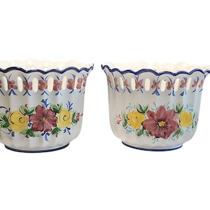 May include: Two white ceramic flower pots with scalloped rims and floral designs. Each pot has a blue rim, cut-out details, and hand-painted flowers in yellow, burgundy, and blue. Ideal for indoor plants.