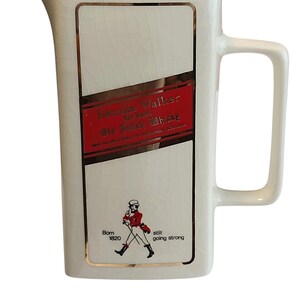 May include: A cream-coloured ceramic jug with a rectangular label featuring the text "Johnnie Walker" and a red and gold design. The jug has a handle and a spout. The bottom of the jug has the text "Born 1820 still going strong".