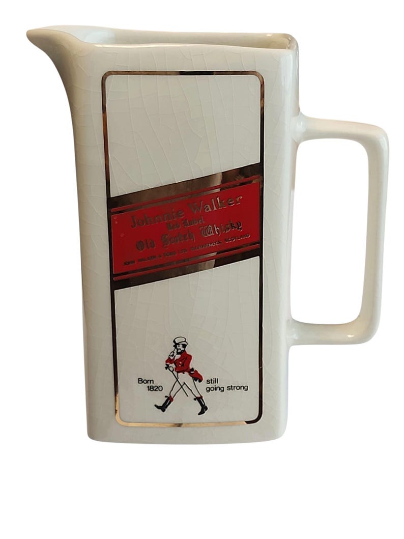May include: A cream-coloured ceramic jug with a rectangular label featuring the text "Johnnie Walker" and a red and gold design. The jug has a handle and a spout. The bottom of the jug has the text "Born 1820 still going strong".