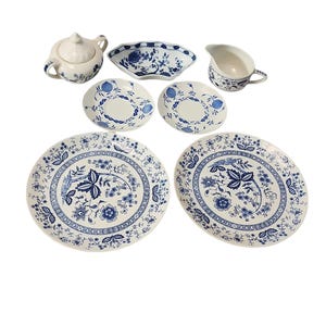 May include: A collection of white porcelain dishware with blue floral patterns. The set includes plates, bowls, a creamer, and a sugar bowl with a lid. The design features detailed botanical motifs and decorative borders.