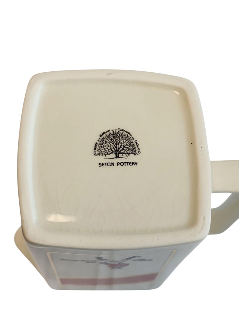May include: Cream ceramic mug with a square base and a handle. The base features a black tree logo and the words "SETON POTTERY". The mug has a rectangular shape and a slightly glossy finish. Made in England.