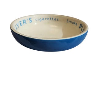 Può includere: Un posacenere in ceramica blu e bianco con la scritta "AYER'S cigarettes Smoke PL". Il posacenere ha una forma arrotondata con esterno blu e interno bianco. Un oggetto vintage.