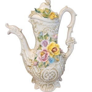 Op de afbeelding: Een witte porseleinen theepot met een bloemenmotief. De theepot heeft een deksel met een bloemenmotief en een handvat. De theepot is versierd met gele, roze en groene bloemen.