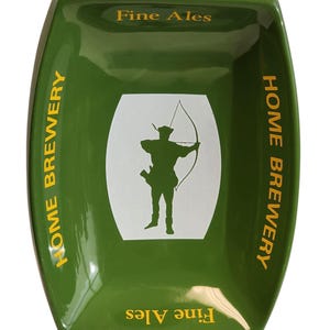 Può includere: Un vassoio rettangolare in ceramica verde con lati arrotondati, caratterizzato da un ovale centrale bianco contenente la sagoma nera di un arciere. Il testo "HOME BREWERY" è stampato in giallo sui lati, con "Fine Ales" in giallo in alto e in basso.