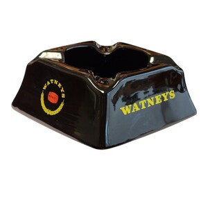 Può includere: Un posacenere quadrato in ceramica marrone scuro con quattro incavi per sigarette. Il lato presenta la scritta gialla WATNEYS e un logo circolare con la parola WATNEYS, un'icona a forma di botte rossa e un disegno di spiga di grano gialla.