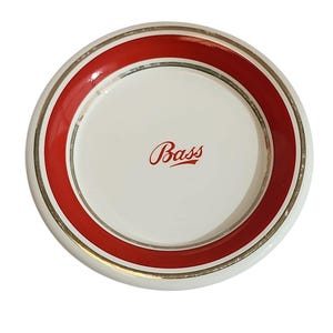 Può includere: Un piattino rotondo in ceramica bianca con un'ampia fascia rossa lungo il bordo e una sottile linea color oro. Al centro del piattino è stampata la parola Bass in un carattere corsivo rosso. Il design è pulito, circolare e simmetrico.