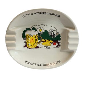 Può includere: Un posacenere ovale in ceramica bianca con il testo THE PINT WITH REAL FLAVOUR stampato due volte lungo il bordo. Il grafico centrale raffigura un boccale di birra giallo con schiuma, una pagnotta, un pezzo di formaggio e un pomodoro rosso.