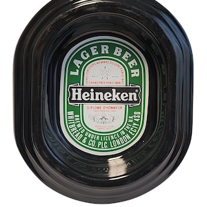 Puede incluir: Un cenicero ovalado de color negro para cerveza Heineken con una etiqueta central verde y blanca. La etiqueta muestra el texto LAGER BEER, Heineken, DIPLOME D'HONNEUR y WHITBREAD & CO. PLC. LONDON EC1Y 4SD, junto con detalles de premios hist&oacute;ricos.