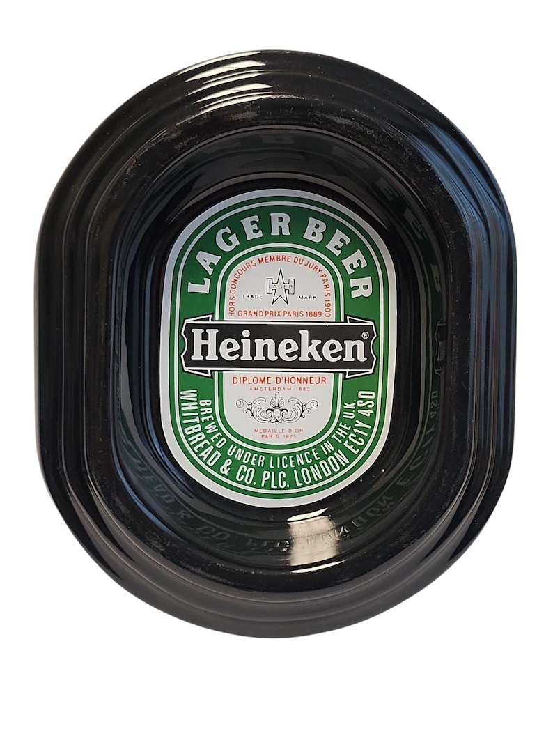 Puede incluir: Un cenicero ovalado de color negro para cerveza Heineken con una etiqueta central verde y blanca. La etiqueta muestra el texto LAGER BEER, Heineken, DIPLOME D'HONNEUR y WHITBREAD & CO. PLC. LONDON EC1Y 4SD, junto con detalles de premios hist&oacute;ricos.