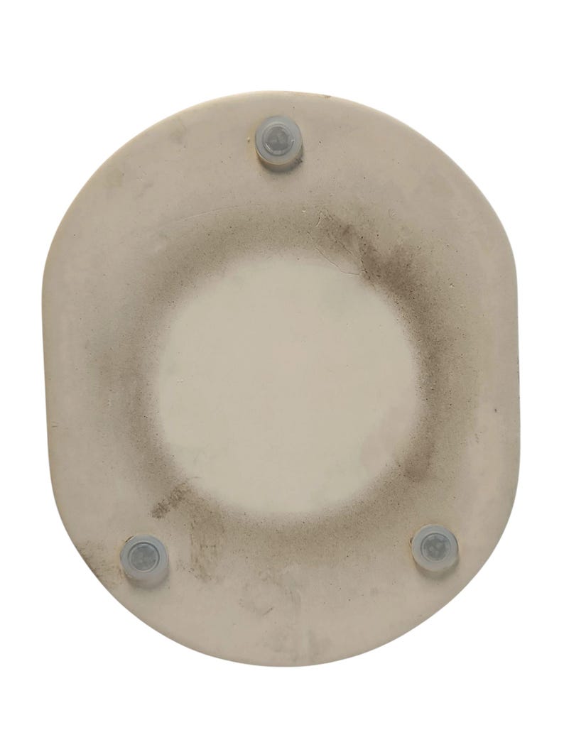 Puede incluir: Un salvamanteles circular de piedra color blanco roto con una superficie central lisa y un borde ligeramente texturizado. La parte inferior incluye tres peque&ntilde;as patas de silicona circulares y transparentes colocadas sim&eacute;tricamente.