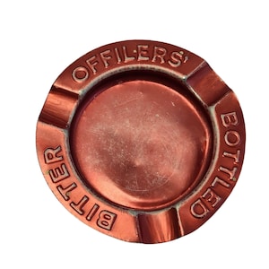 Può includere: Un posacenere rotondo color rame metallizzato con tre tacche lungo il bordo. La scritta in rilievo "OFFILERS' BOTTLED BITTER" è impressa lungo il bordo in un motivo circolare. La parte centrale presenta segni di usura superficiale.