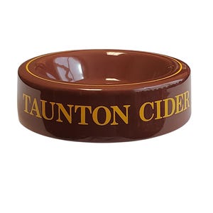 Può includere: Un posacenere rotondo in ceramica marrone con finitura lucida. Il testo TAUNTON CIDER è stampato in lettere maiuscole gialle sul lato. Una sottile linea circolare gialla è visibile lungo il bordo interno della ciotola dal centro concavo.