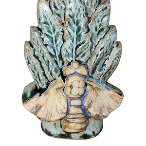 Puede incluir: Un jarrón de cerámica con un esmalte verde y marrón. El jarrón tiene forma de abeja con alas y cuerpo. La abeja está rodeada de hojas.