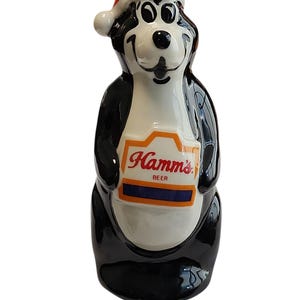 Può includere: Una statuetta in ceramica di un orso bianco e nero che indossa un cappello da Babbo Natale rosso. L'orso ha un petto bianco con un cartello bordato di arancione che riporta la scritta rossa Hamm's e la parola BEER sopra una striscia blu scuro.