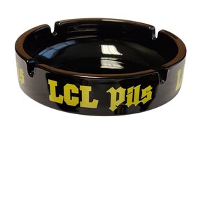 Può includere: Un posacenere rotondo in ceramica nera con la scritta LCL Pils stampata in lettere gialle sul lato esterno. Il posacenere ha una finitura liscia e lucida e include quattro tacche distanziate uniformemente lungo il bordo superiore.