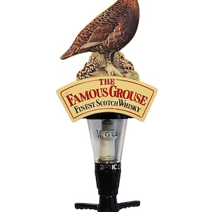 Può includere: Un dosatore per alcolici da bar con la sagoma di un gallo cedrone marrone appollaiato su una roccia. Sotto l'uccello, un cartello color crema reca la scritta THE FAMOUS GROUSE FINEST SCOTCH WHISKY. Il misuratore trasparente riporta 1/6 GILL e OPTIC.