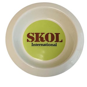 Può includere: Una ciotola rotonda in ceramica bianca con una grafica circolare verde lime al centro. Il testo SKOL è stampato in grandi lettere marroni con grazie, con la parola International scritta in un carattere più piccolo direttamente sotto il marchio.