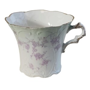 Puede incluir: Una taza de té blanca con un borde festoneado y un asa. La taza presenta un exterior verde pálido con delicados patrones florales morados. El borde y el asa están acentuados con ribetes dorados, agregando un toque de elegancia al diseño.